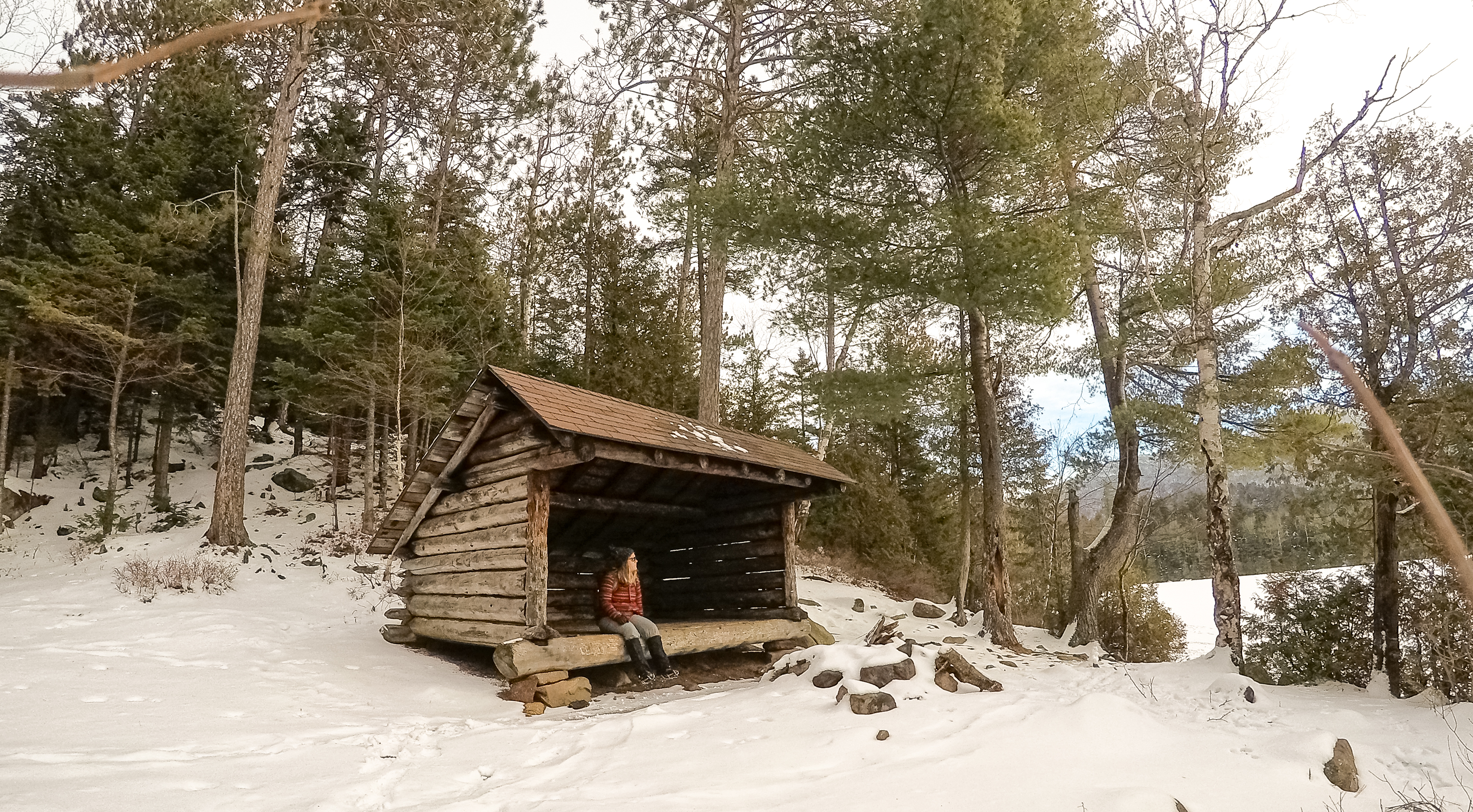 A woman in a red jacket sits in a lean-to next to a frozen pond covered in snow.
