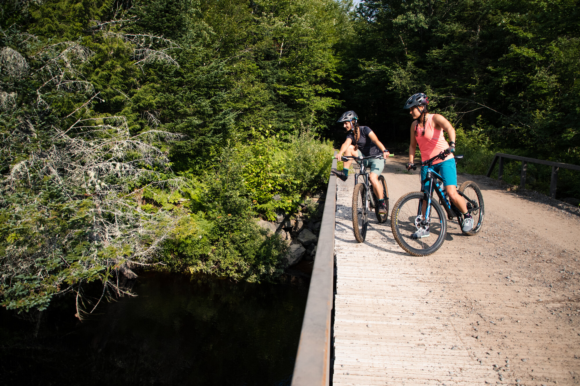 Two women on bikes stop on a dirt bridge to look at the stream below. 