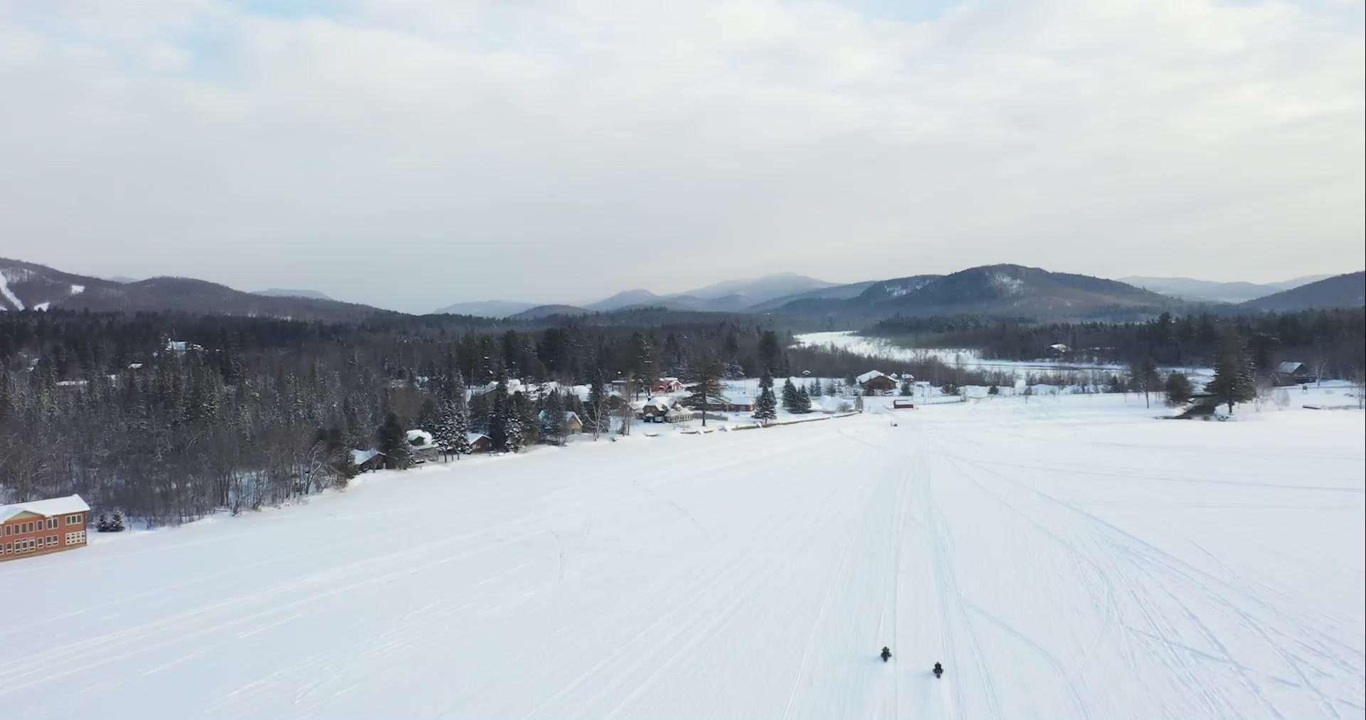 An aerial view of two snowmobilers riding over a snow-covered frozen lake towards the mountains