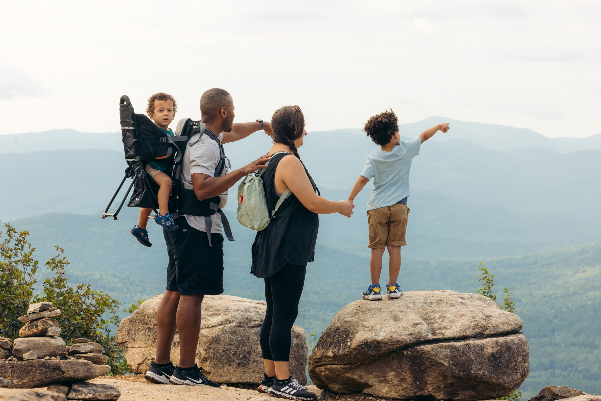 Family of four stands on summit looking at view with youngest child in backpack carrier