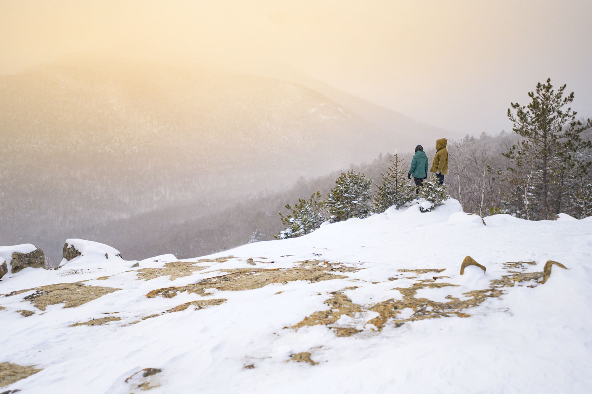 Two hikers wearing winter gear and snowshoes stand on Cobble Lookout, with scenic mountains in the distance