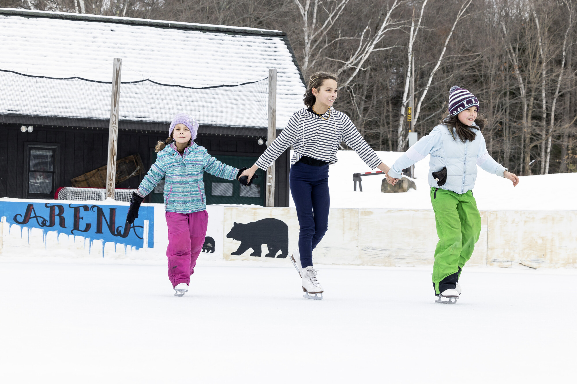 Two younger kids hold hands with an older kid as they all skate on the outdoor ice rink at Mt Sabattis Recreation Area in Long Lake