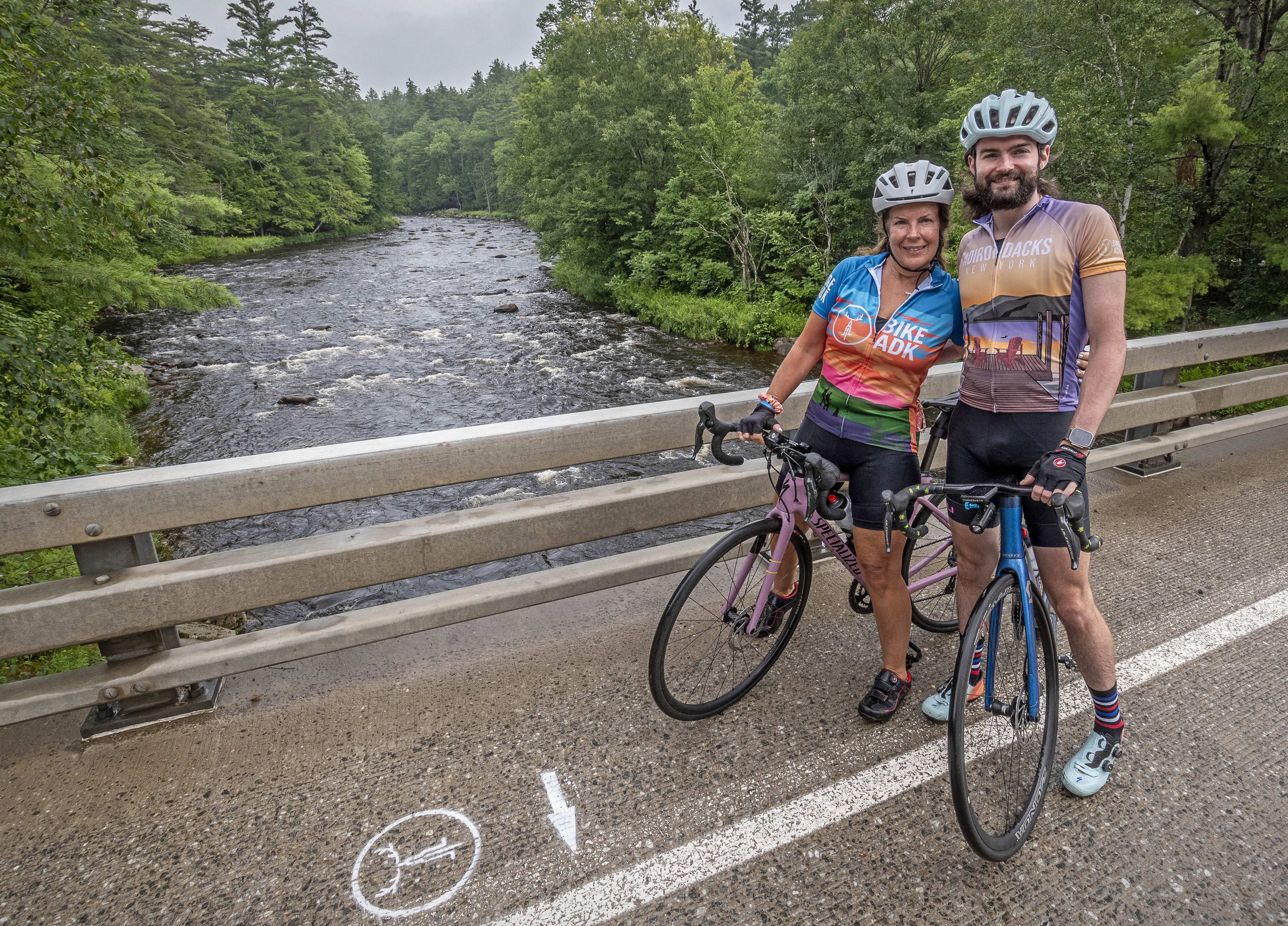 Two riders poses on their bikes in front of a river. 