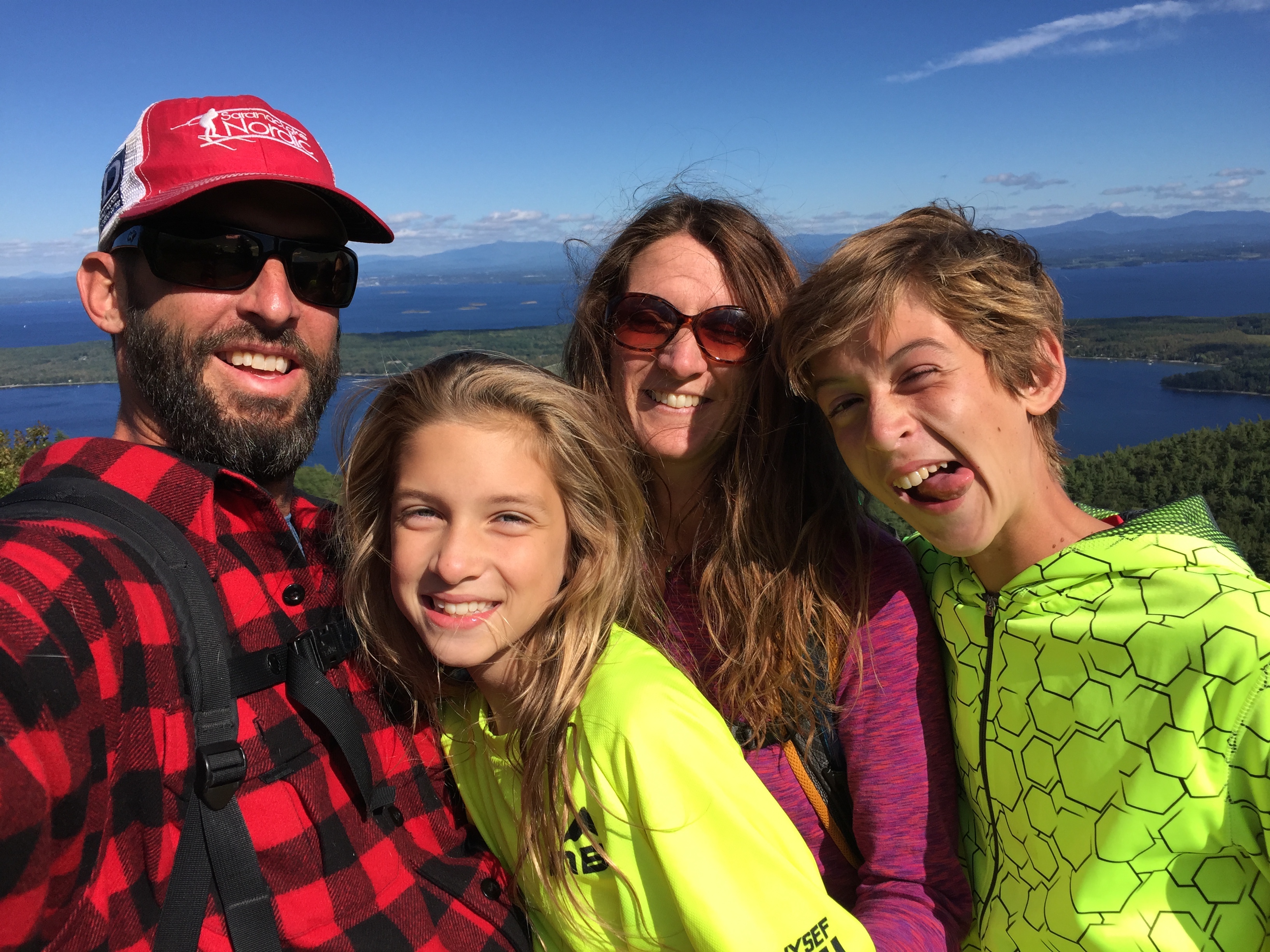 A man and woman with their two children smile while standing on top of a mountain with water and the High Peaks behind them