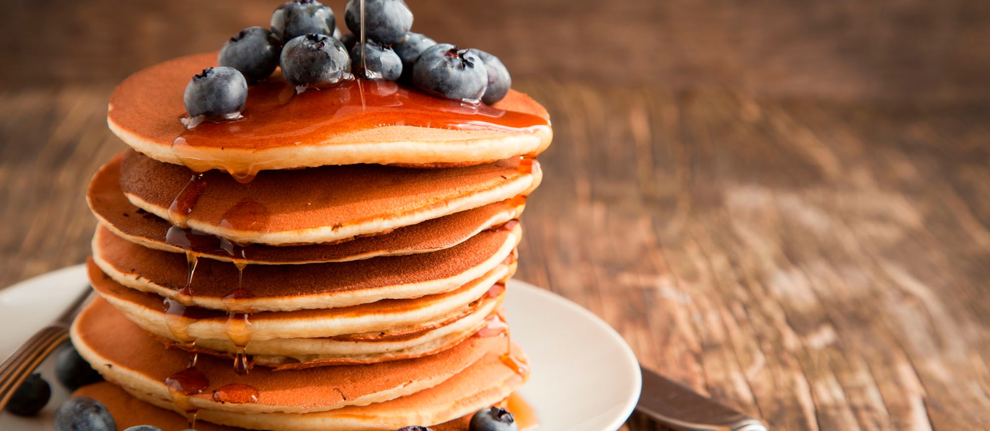 Maple syrup is poured over a stack of pancakes topped with fresh blueberries