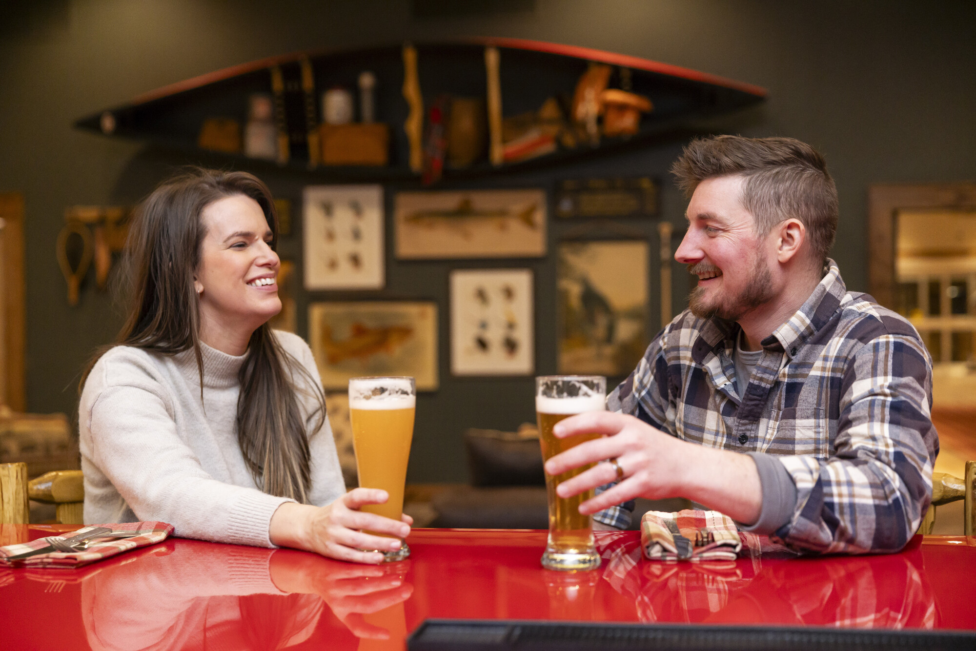 A man and woman sit at a wood-topped bar, drinking glasses of beer. Rustic Adirondack decor is visible in the background.