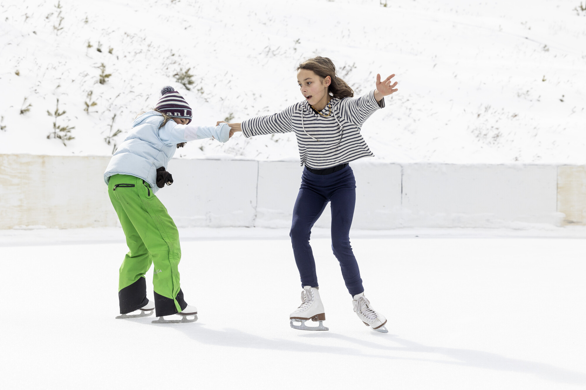 two young girls in warm clothes ice skate on an outdoor rink. 