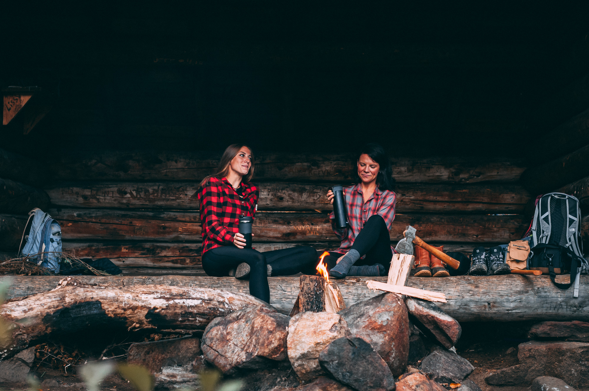 Two women relax in a lean-to with hot drinks and flannel shirts 