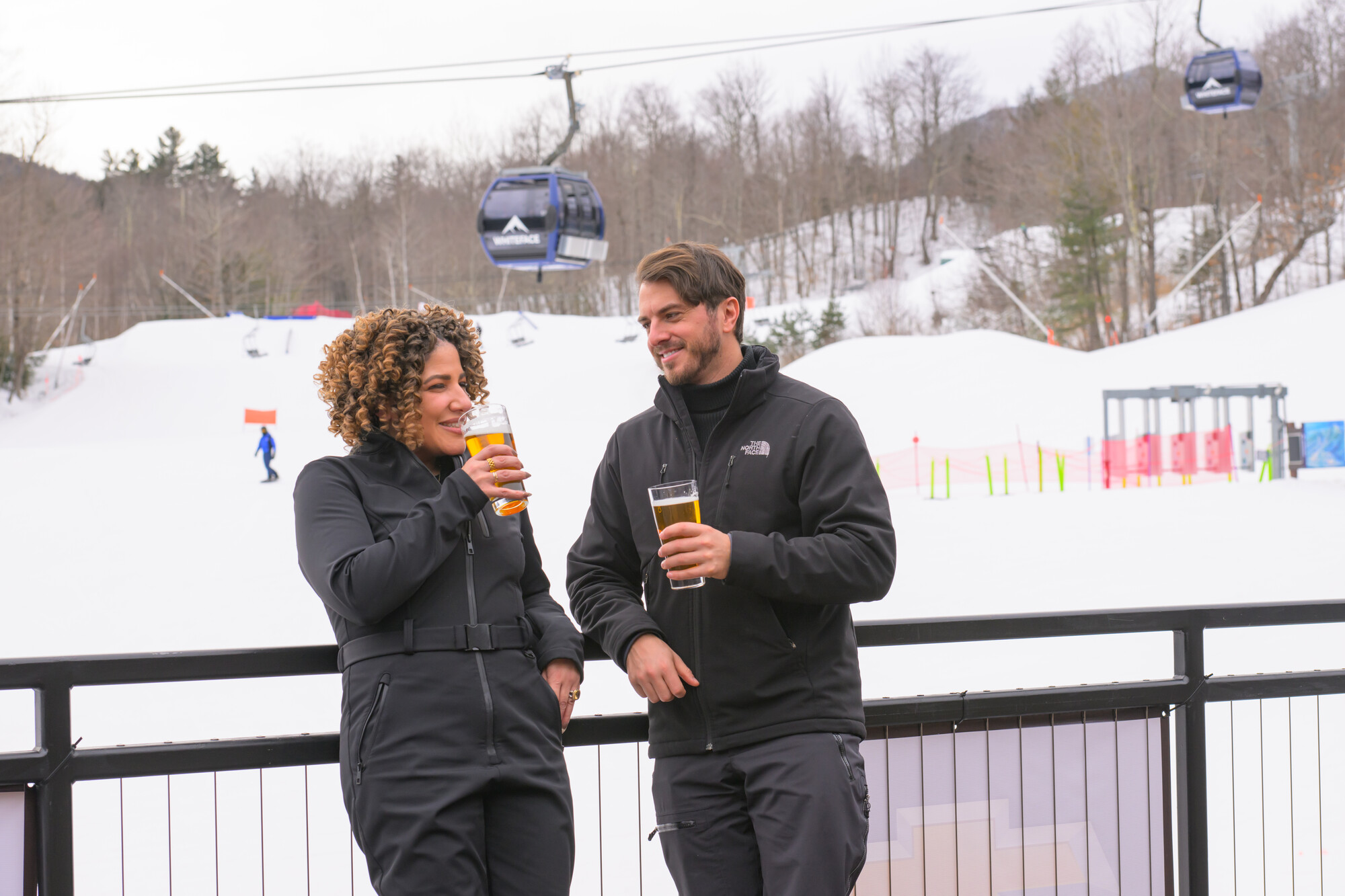 A couple drinking beers on the deck of Whiteface base lodge with the Cloudsplitter Gondola behind them.