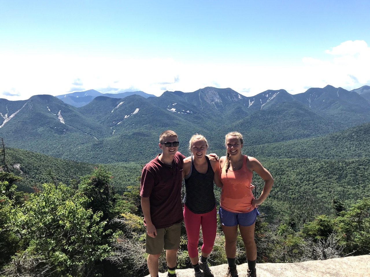 A trio of people stand on a mountain summit with large mountains in the background on a sunny summer day.