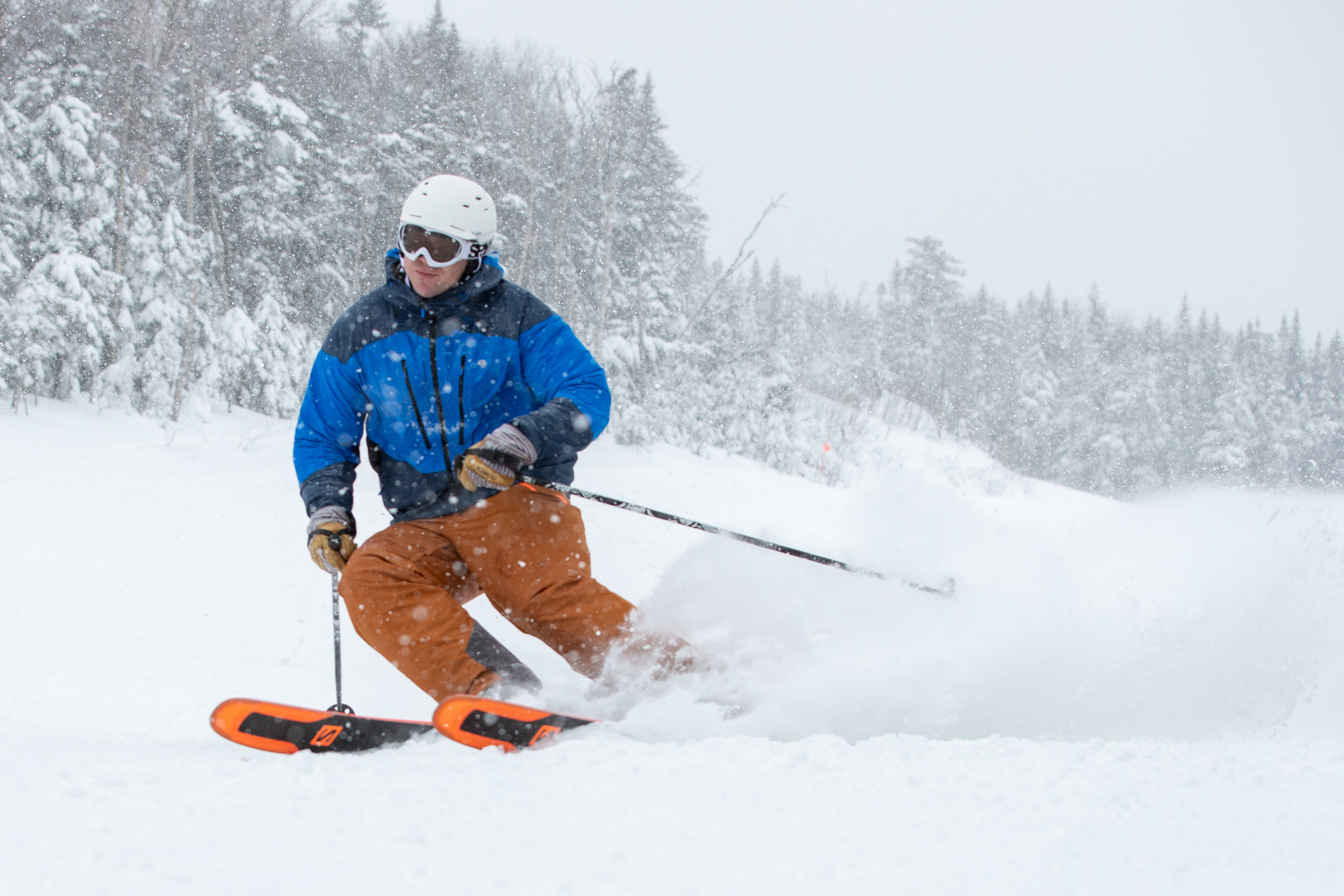 A skier in a blue coat and orange pants skis downhill during a snow storm.