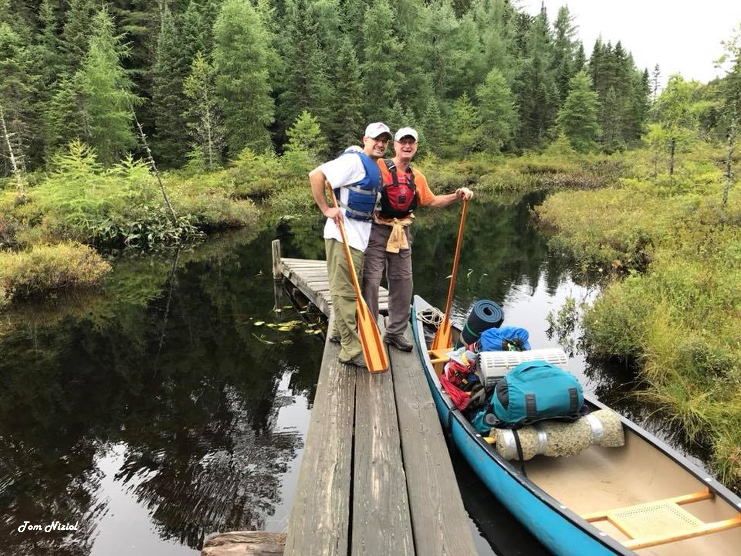 Two men stand smiling next to a packed canoe ready to go camping.