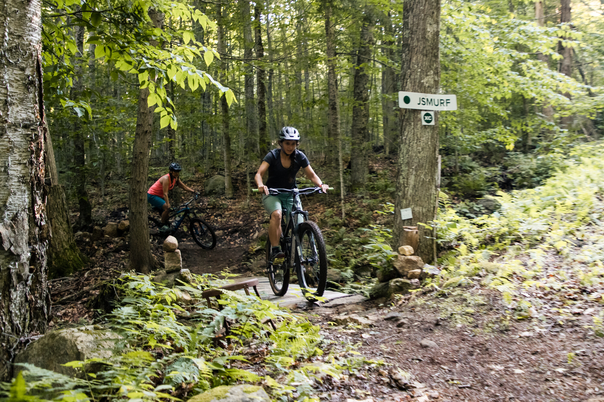 Two women on mountain bikes riding past a trail sign on a tree in a bright forest.