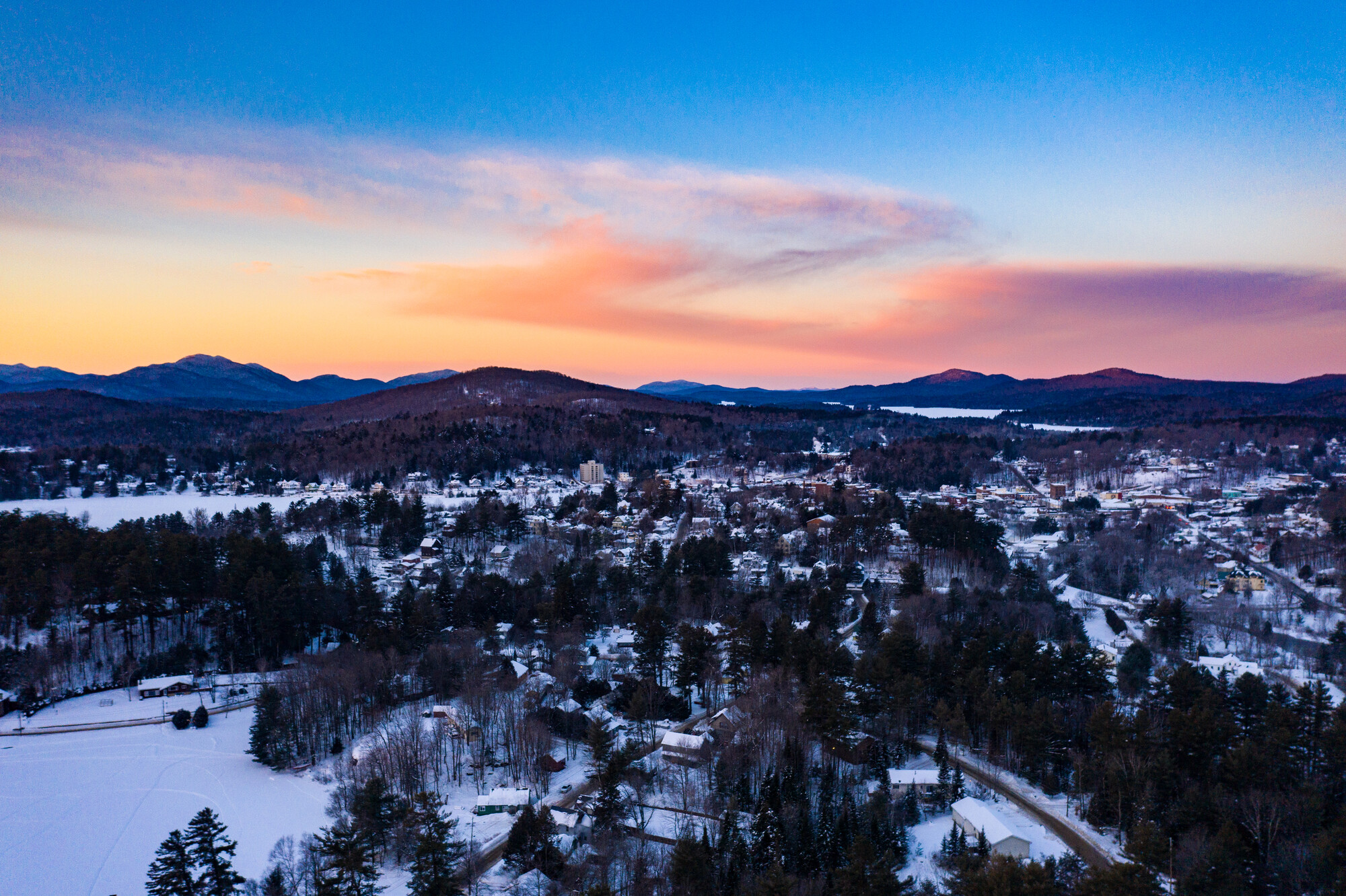 The sun sets behind a snow-covered town with mountains in the background.