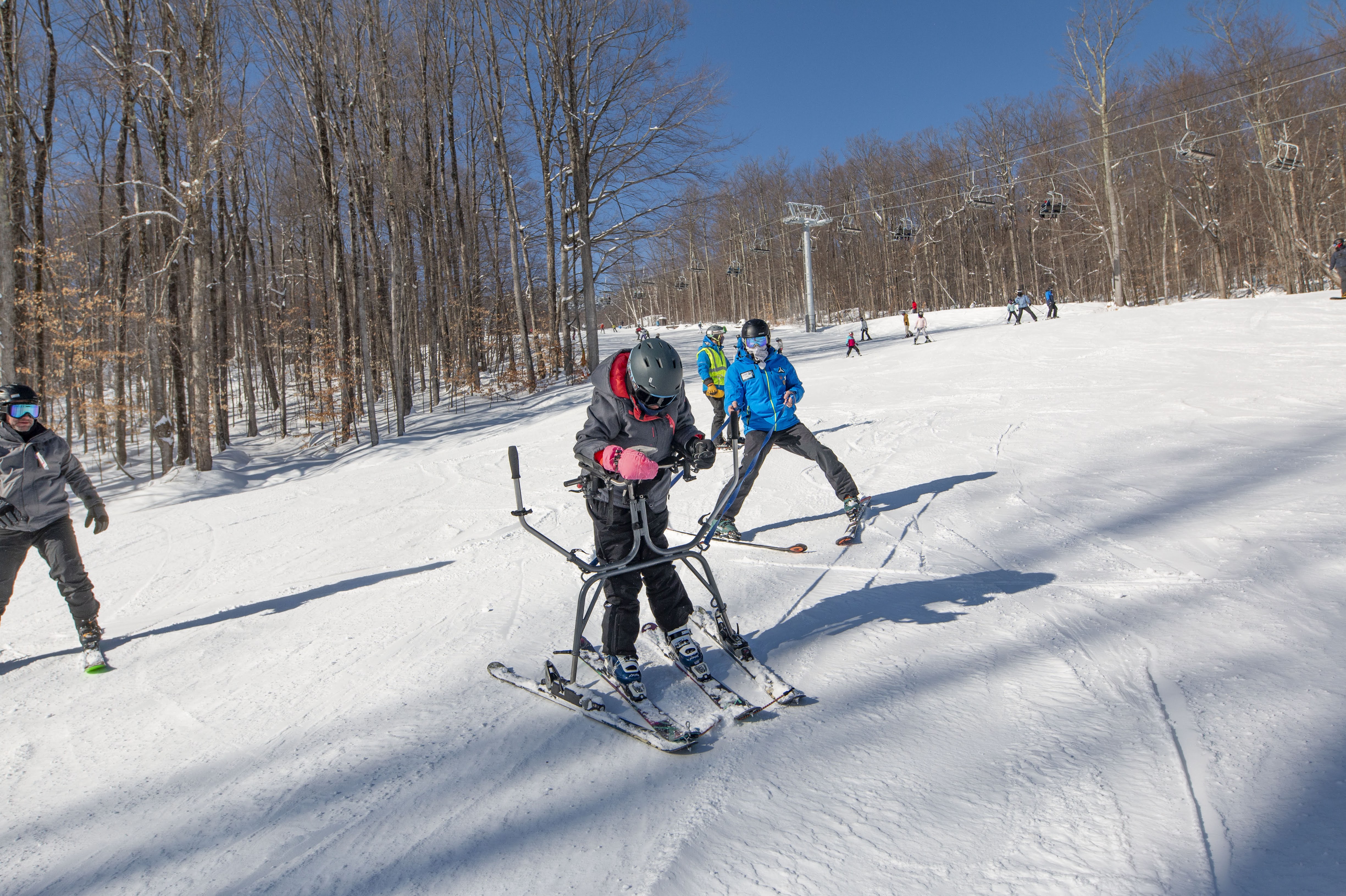 Someone in a blue coat gives an adaptive, para ski lesson to someone who uses outriggers for support.