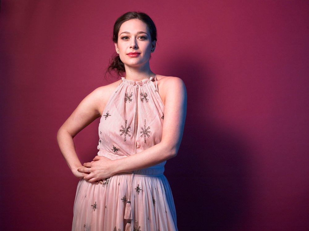 A dark-haired woman in a pink dress stands in front of a maroon wall in a portrait.
