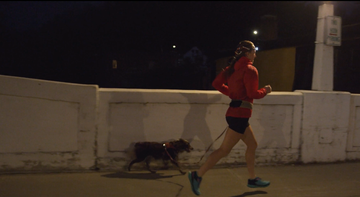 A small dog runs on a sidewalk with its owner before dawn
