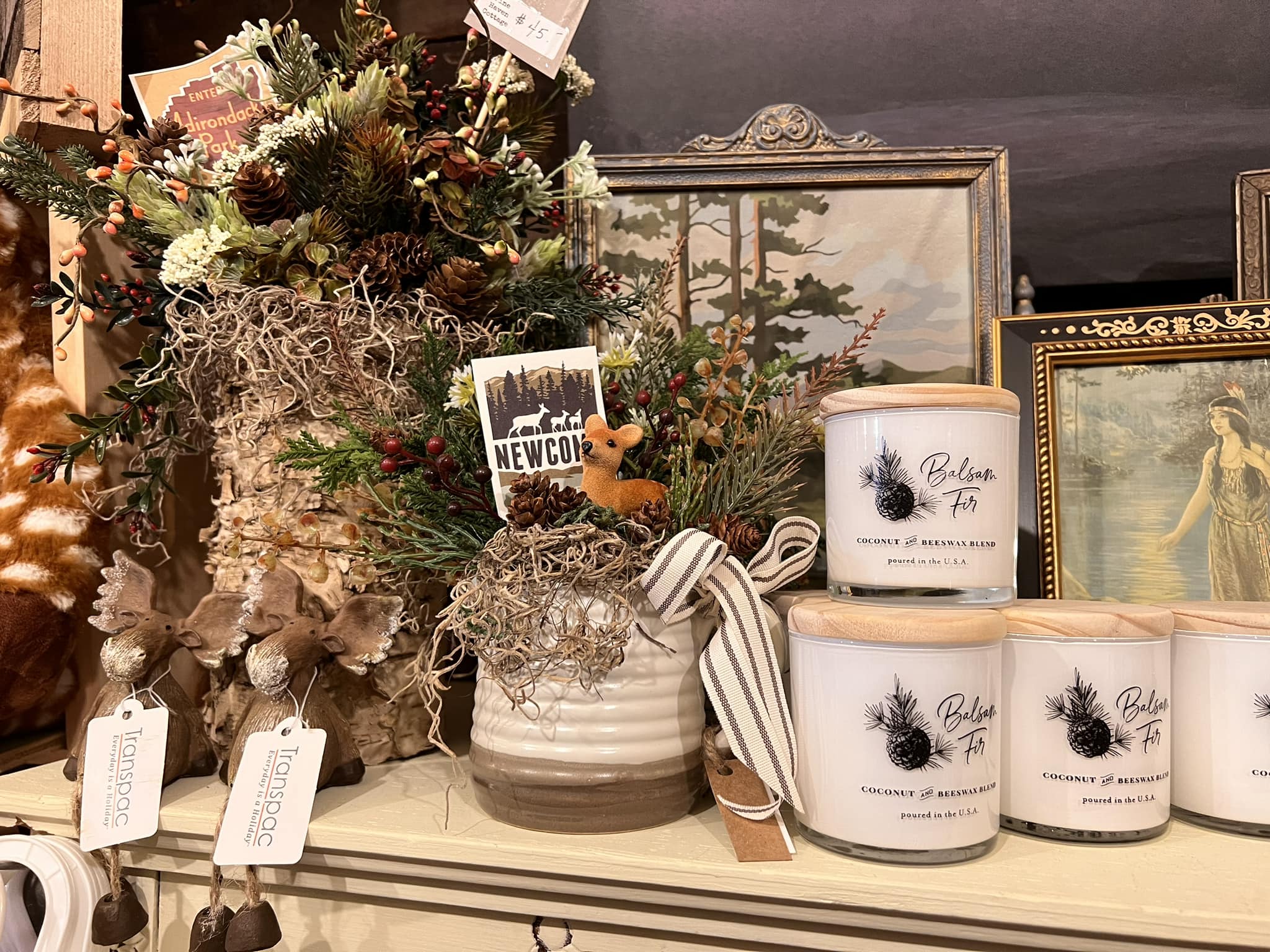 Pine boughs and cones decorate a shelf with candles in glass jars and prints in vintage wood frames.