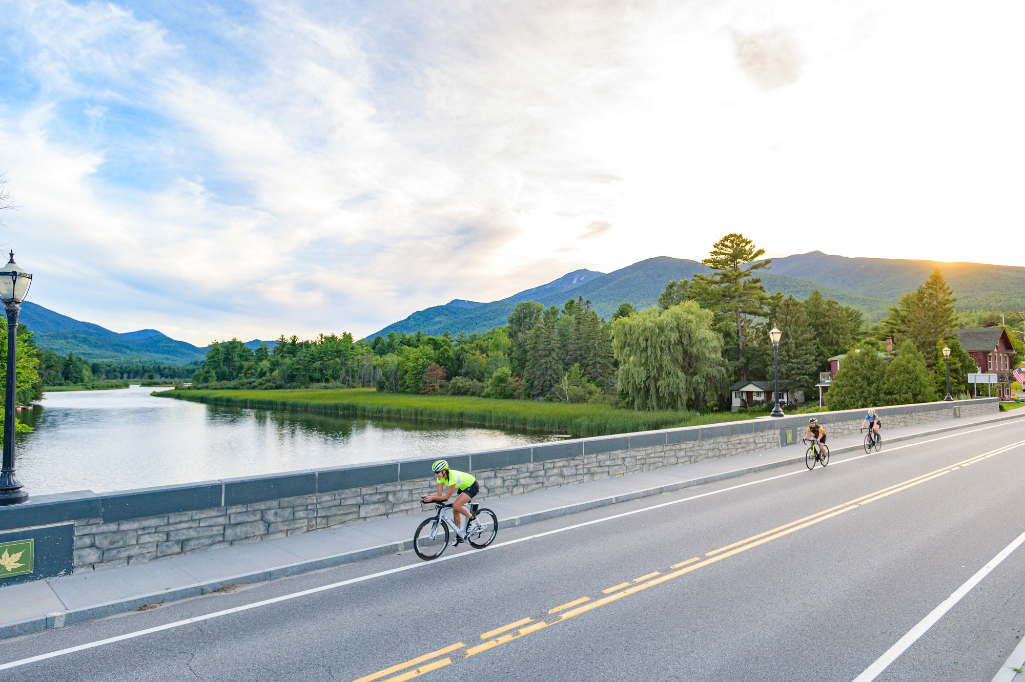 Three cyclists passing over a stone bridge with mountains and sunset in the background.