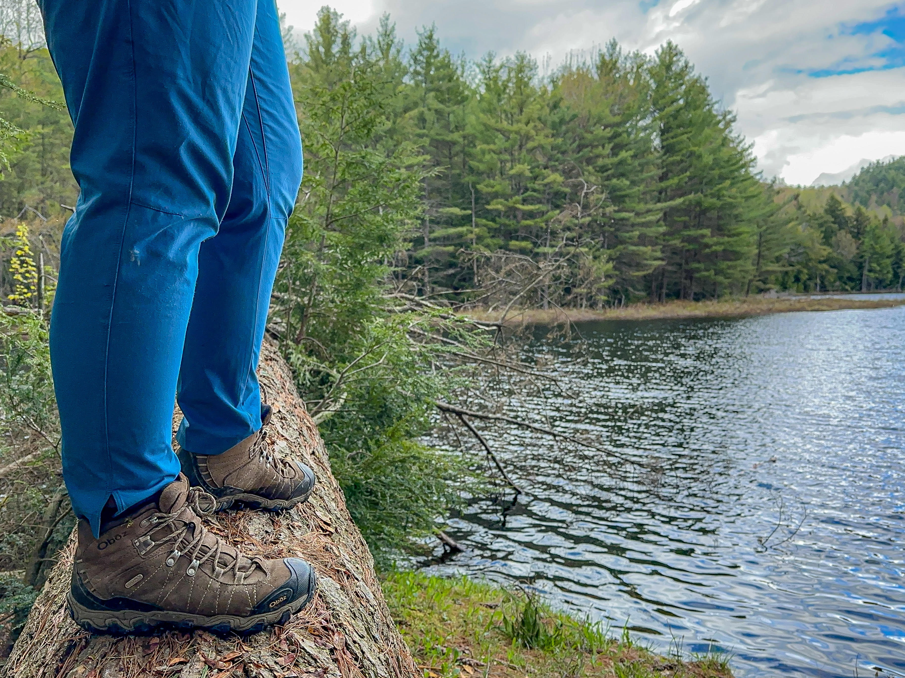 Someone with blue pants and hiking boots standing on a log, only the lower parts of their legs showing, next to a pond.