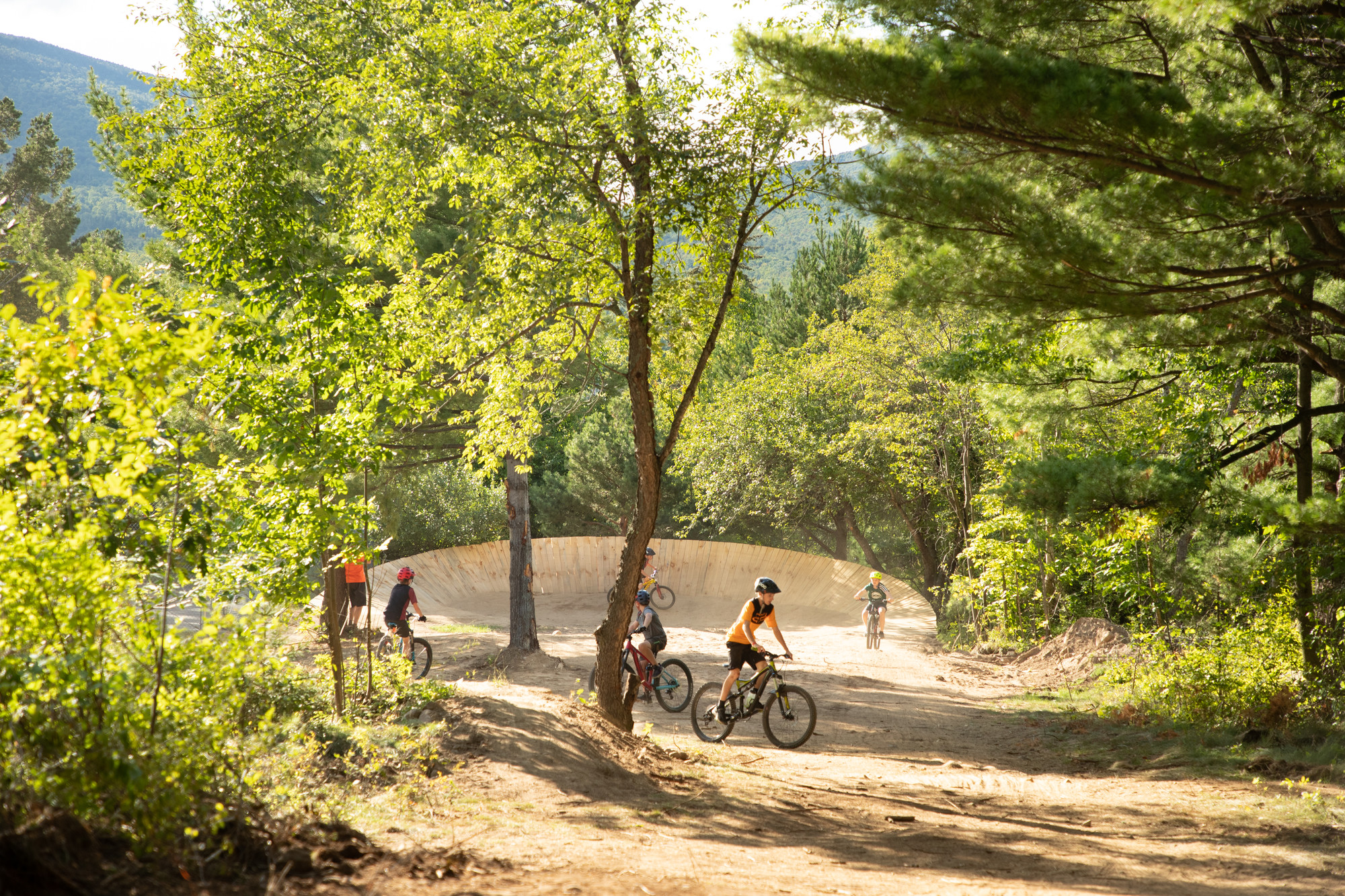 Kids ride around bike park with features and trees on sunny day