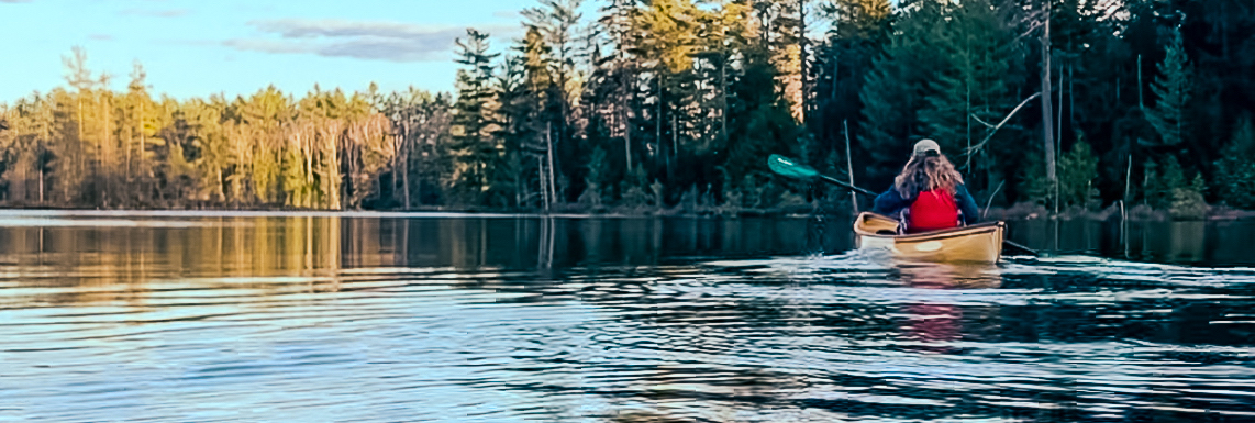 A woman paddling a solo canoe with a red life vest on on a pond at sunset