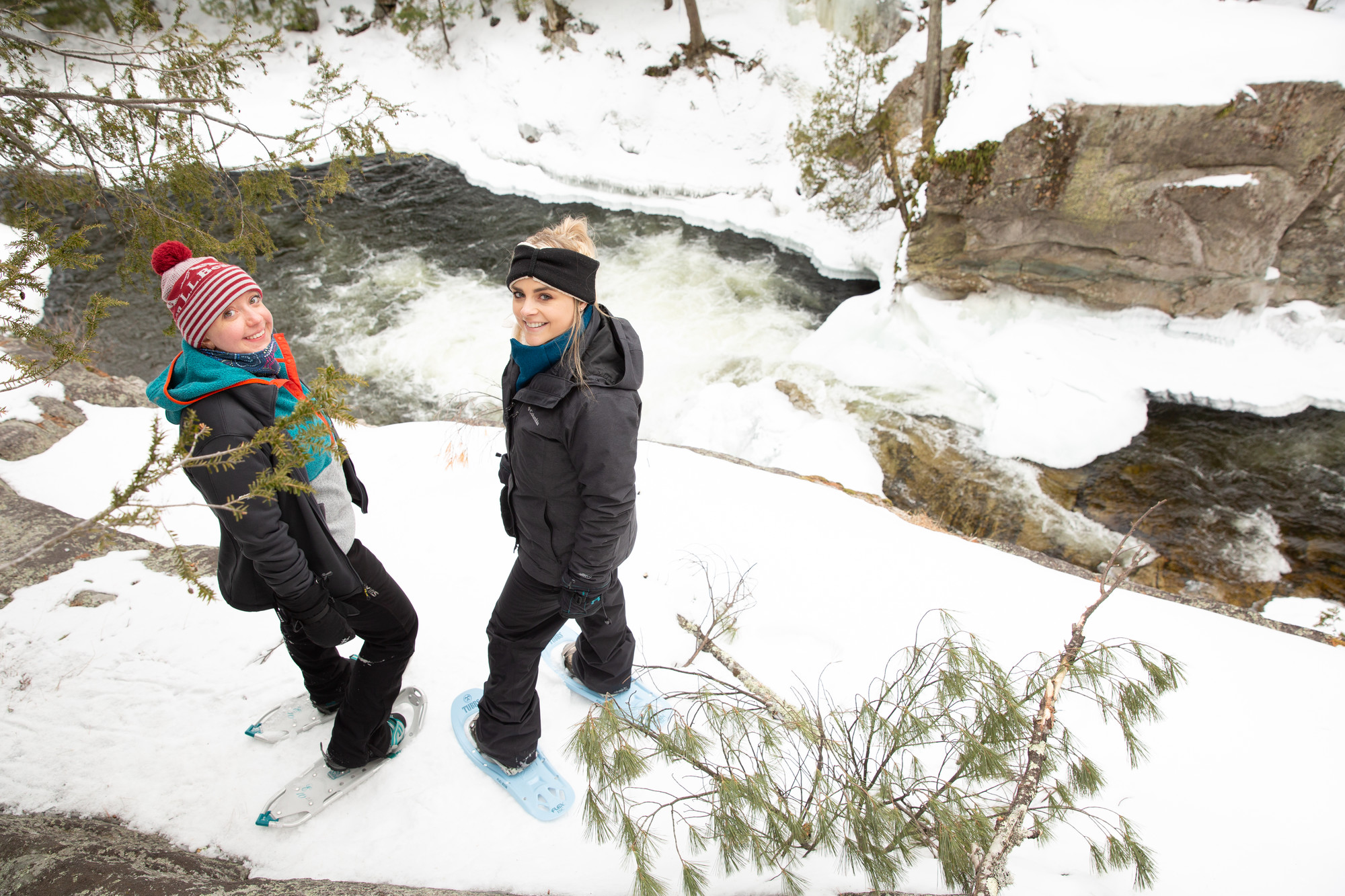 Two women look up at the camera from a snowy ledge above an icy river, while on snowshoes.