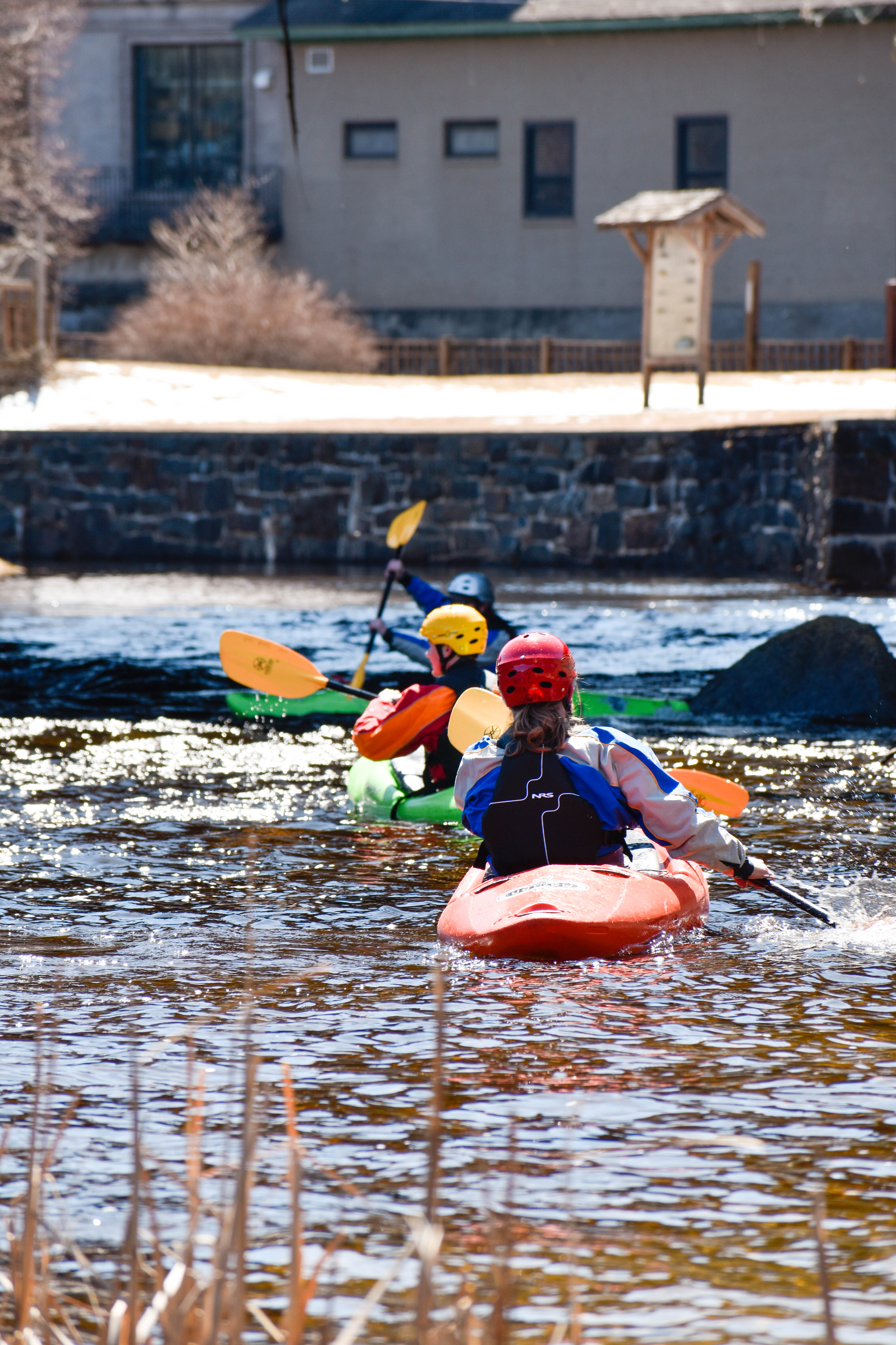 Kayakers paddle together down a river with a town's skyline in the background.