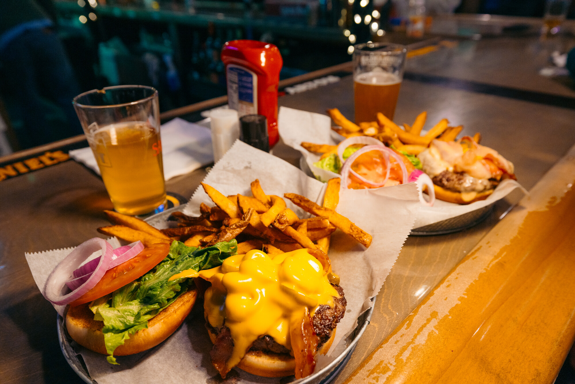 Two plates of cheese burgers and fries with beer on the side. 