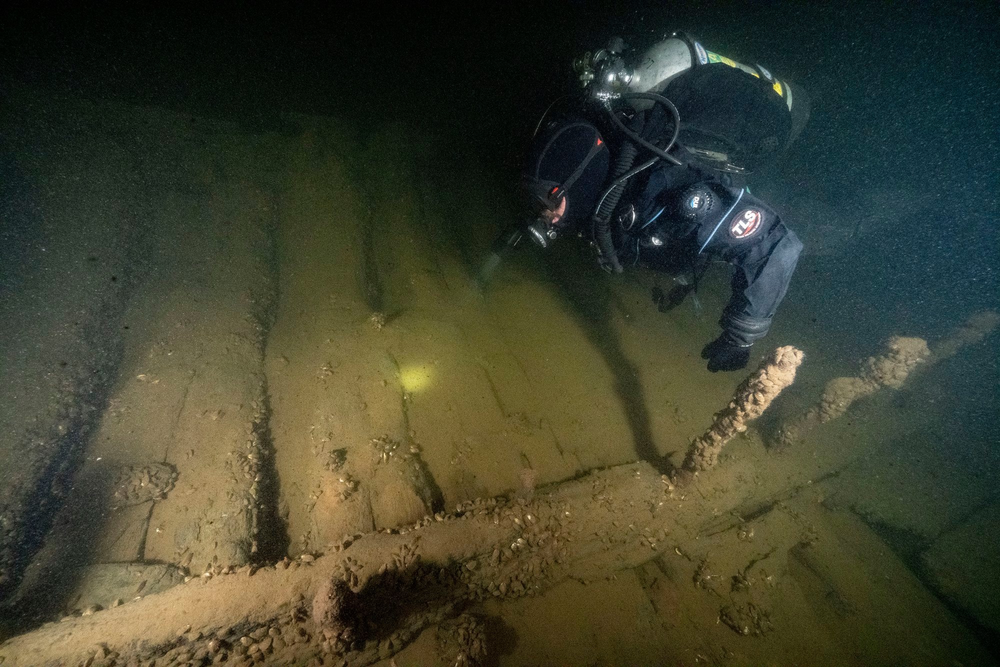 A scuba diver floats above the wooden remains of a boat in dark water. 