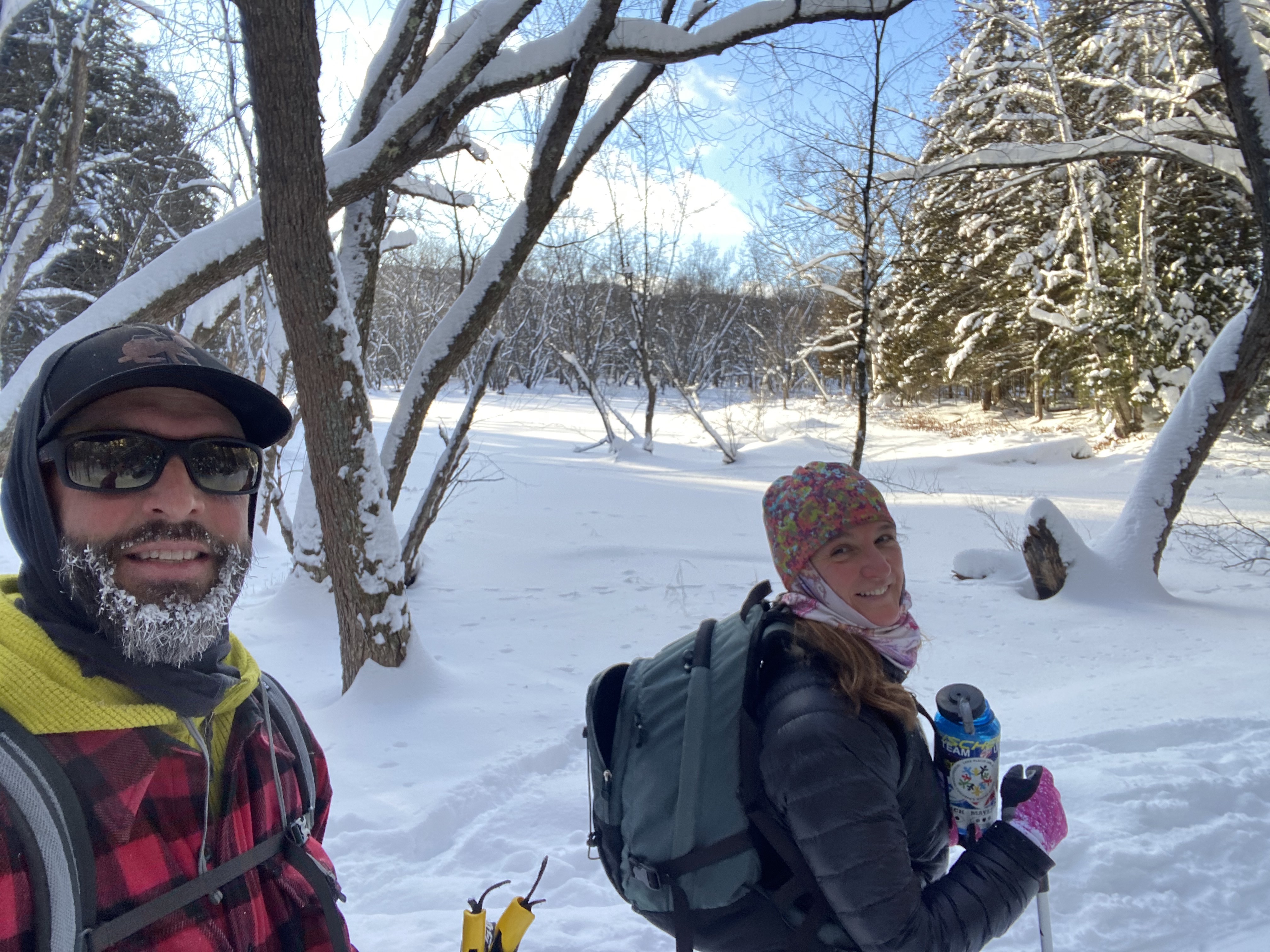 A man with a beard covered in frost and a woman smile for a photo while walking in the snow