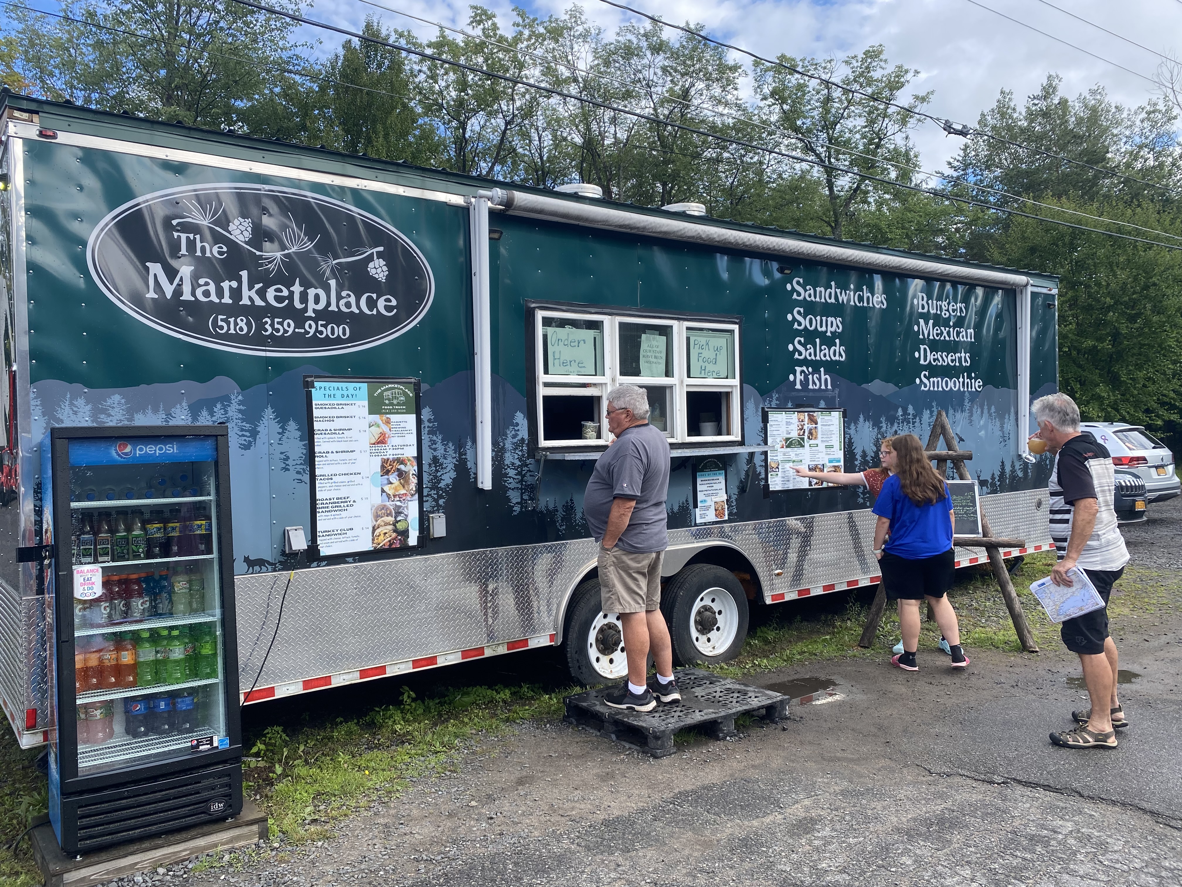 A large green food truck advertising its menu; people wait in line at the window.