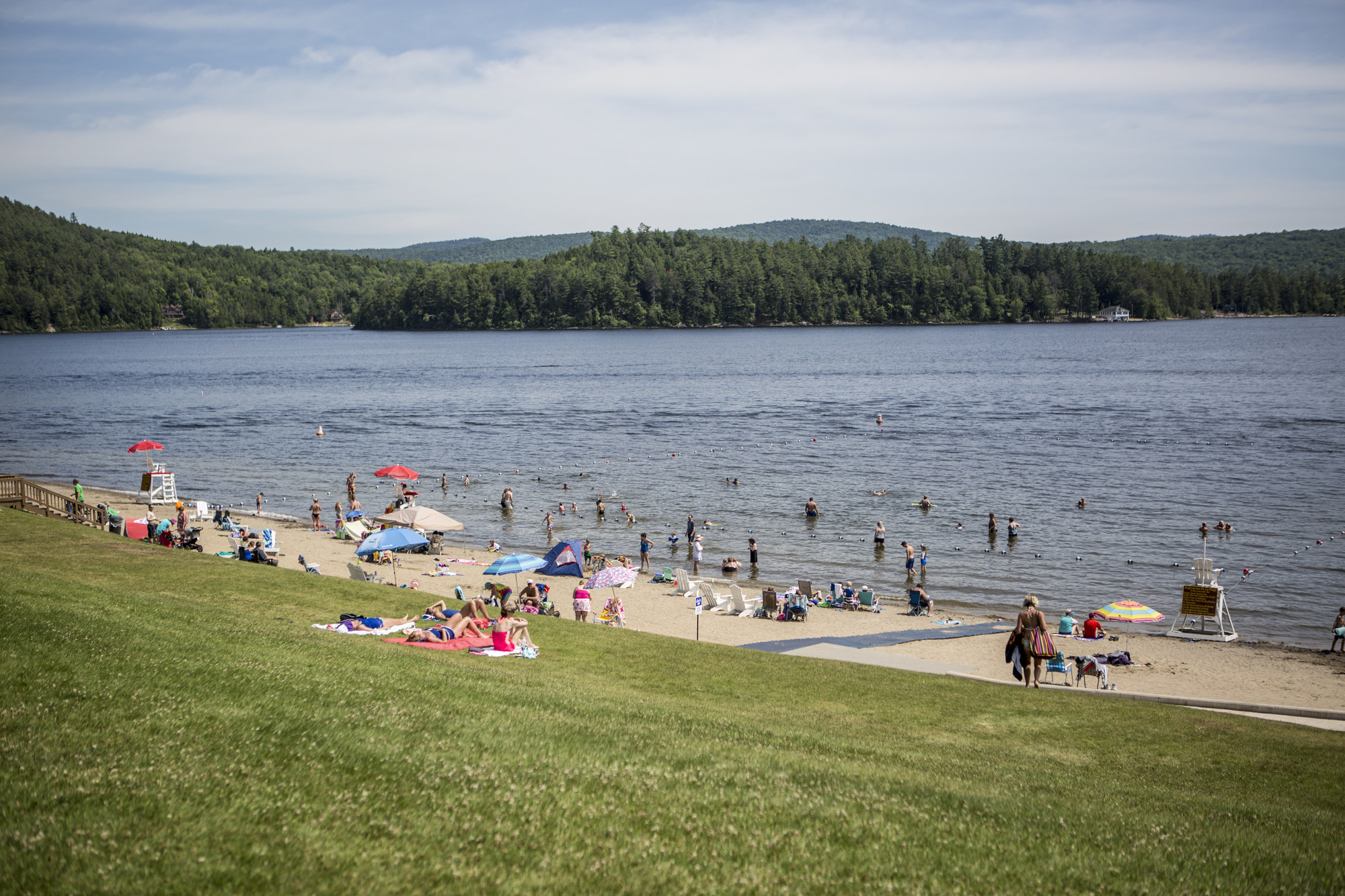 A wide view of people lounging and playing on a sandy beach, while others swim in a broad, blue lake.