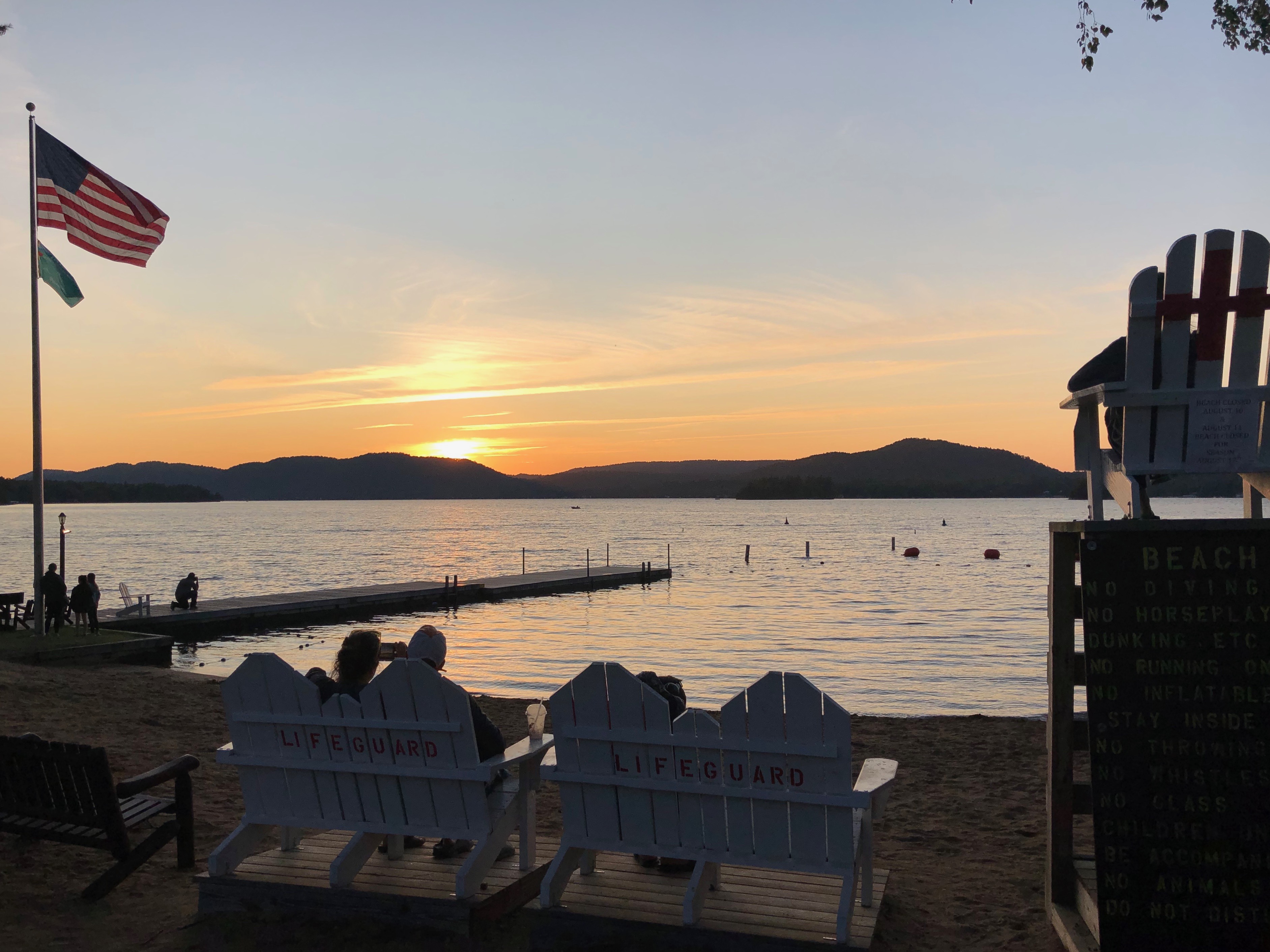 A view of a beach with a sunset over the lake and mountains beyond.